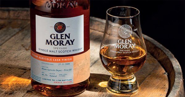 eine Flasche Glen Moray Elgin Curiosity 2006 Rhum Agricole Cask Finish nebst einem Glas auf einem Holzfass