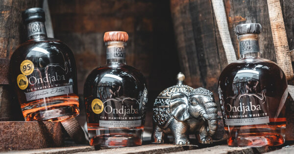 Ondjaba Namibian Whiskey drei Flaschen Whiskey und ein Metallelefant stehen auf einer Palette, umgeben von Holzfässern