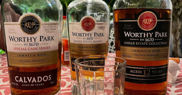 von links nach rechts aufgereit: eine Flasche Worthy Park Calvados, eine Flasche Worthy Park Single Estate Reserve und eine Flasche Worthy Park Single Estate 12 Jahre. Der Fokus liegt auf den Etiketten. Im Vordergrund ein Glas