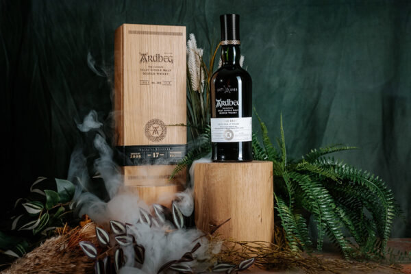 das Foto zeigt eine Flasche Ardbeg Single Cask No. 385 2008/2025 auf einem Holzzylinder vor einem Farn. Daneben eine Holzkiste, davor steigt Rauch auf