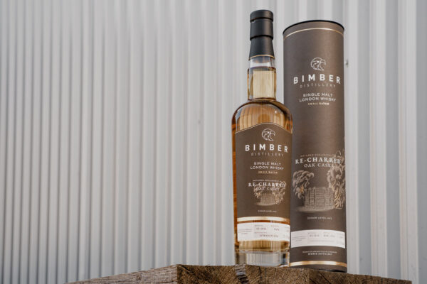 Bimber Re-Charred Oak Cask Batch 2 eine Flasche Bimber Re-Charred Oak Cask Batch 2 nebst Geschenkhülle steht auf Holzbrettern vor einer Halle