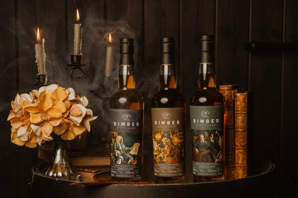 drei Flaschen Whisky stehen samt Büchern, Kerzenständer und Blumenstrauß auf einem Holzfass