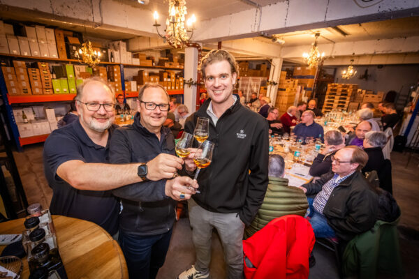 das Foto zeigt Menschen in einer Halle. Zur Kamera gerichtet sind drei Männer, die mit Gläsern anstoßen
