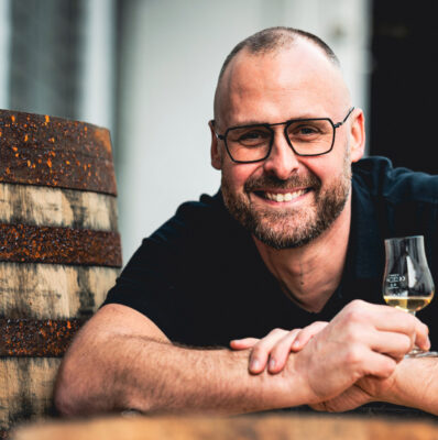 Mario Rudolf ein Mensch mit einem Glas in der Hand lehnt auf ein Holzfass und guckt in die Kamera