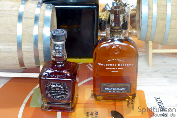 das Foto zeigt zwei Flaschen Whiskey auf einem Holztisch, im Hintergrund zwei kleine Holzfässer
