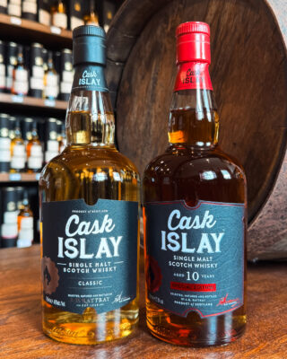 Cask Islay 10 Jahre Sherry Matured zwei Flaschen Whisky auf einem Tisch, im Hintergrund ein Holzfass und ein Regal