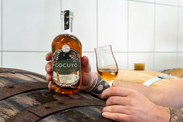 Cocuyo Cask Finished Rum jemand lehnt mit einer Flasche Cocuyo Cask Finished Rum und einem Glas auf einem Fass