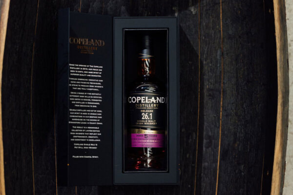 das Foto zeigt eine Flasche Copeland 26.1 Release Single Malt in einer Geschenkverpackung, liegend in einem halbierten Holzfass