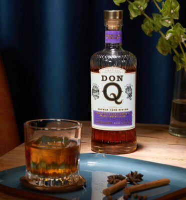 Don Q Double Aged Pedro Ximénez Cask Finish eine Flasche Don Q Double Aged Pedro Ximénez Cask Finish auf einem Holztisch. Vor ihr ein Drink und Gewürze auf einem Teller