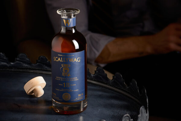 eine geöffnete Flasche Scallywag 18 Jahre auf einem Metalltisch. Im Hintergrund sitzt ein Mann