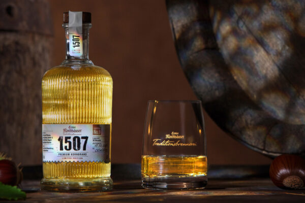 eine Flasche Echter Nordhäuser 1507 Edition No. 9 neben einem Glas. Das Ganze steht auf einem Fass. Im Hintergrund ein weiteres Fass. Links und rechts liegen Kastanien