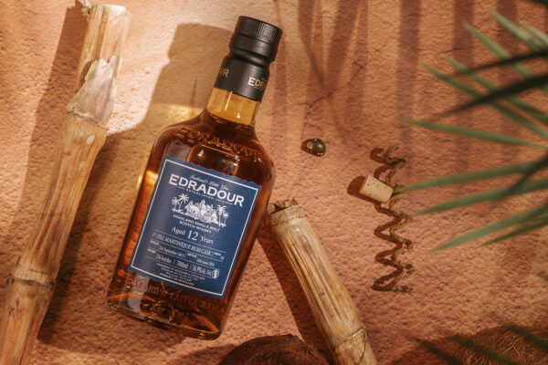 Edradour 12 Jahre Martinique Rum Cask eine Flasche Edradour 12 Jahre Martinique Rum Cask liegt auf einem Sandboden, daneben liegt Bambus