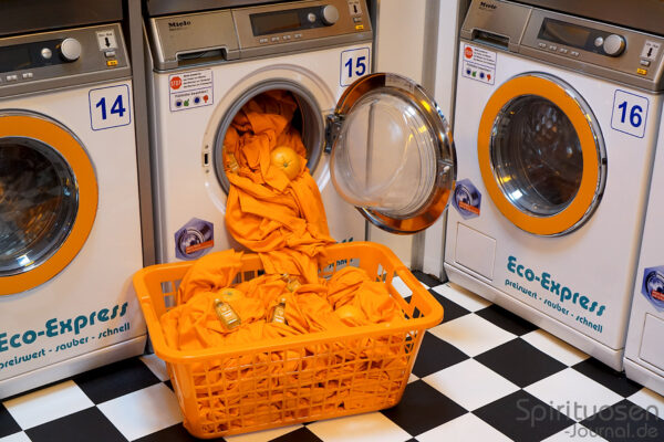 das Foto zeigt eine offene Waschmaschine. Heraus ragen orangen Handtücher. In einem Wäschekorb davor liegen weitere Handtücher, Orangen und Miniaturen Jägermeister Orange
