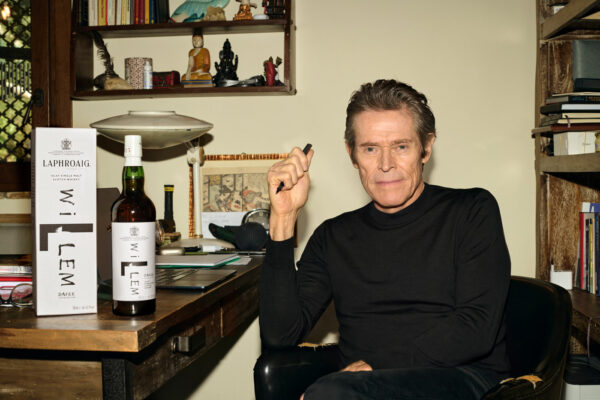 das Foto zeigt Willem Dafoe in einem Büro, auf dem Tisch eine Flasche Whisky