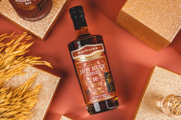eine Flasche MacNair’s Lum Reek 10 Jahre Cask Strength liegt diagonal auf einem roten Tisch. Goldfarbene Klötze stehen drumherum
