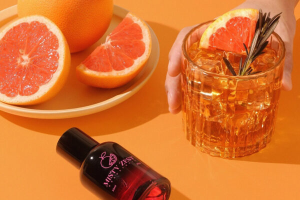 das Foto zeigt einen orangenen Drink, garniert mit Rosmarin und Grapefruit auf orangenem Grund. Davor liegt eine Sprühflasche Misty Zest Grapefruit