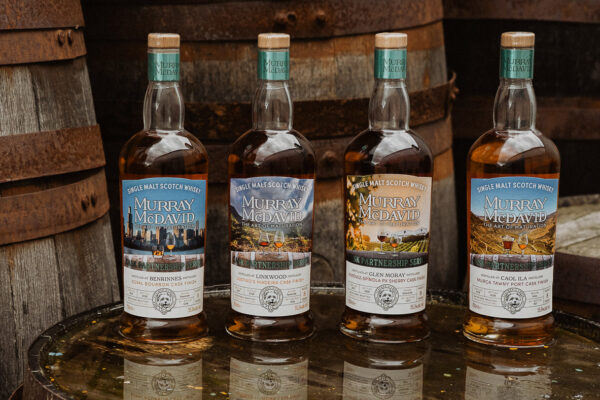Murray McDavid Cask Partnership Series vier Flaschen Whisky auf einem Holzfass. Drum herum weitere Holzfässer