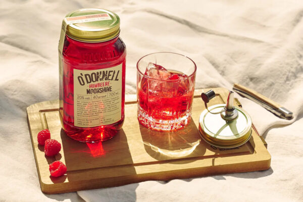 O'Donnell Moonshine Himbeere das Foto zeigt ein Einmachglas O'Donnell Moonshine Himbeere nebst Drink auf einem Holzbrett auf einem Lacken in der Sonne