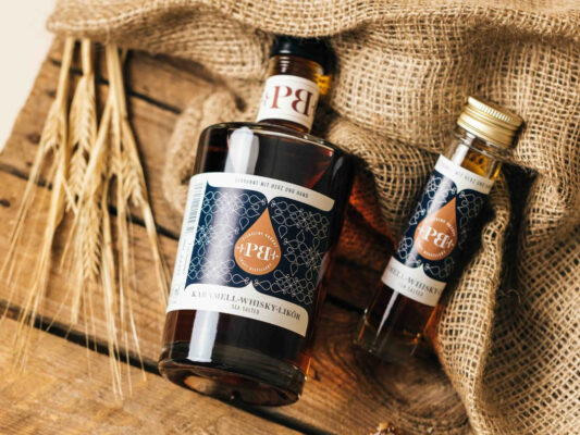 Piekfeine Brände Karamell-Whisky-Likör Sea Salted eine große und eine kleine Flasche Piekfeine Brände Karamell-Whisky-Likör Sea Salted liegen auf einem Sack auf einer Holzpalette