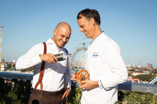 Nicolas Kröger von Wagemut und Magnus Müller-Rischart von Rischart ein Mensch schenkt einem anderen, der eine Brezel in der Hand hält, Gin in ein Glas ein