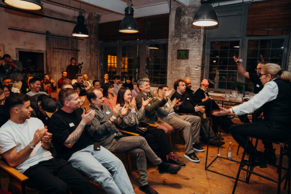 das Foto zeigt Menschen, sitzend in einem alten Industriegebäude. Vorne sprechen zwei Menschen