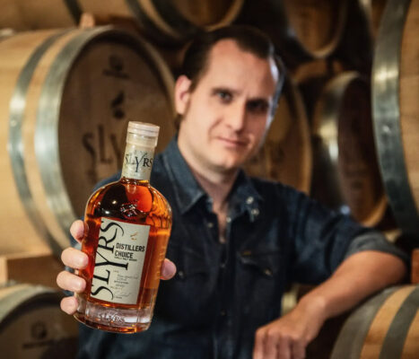 Slyrs Distillers Choice Rhum Agricole Cask ein Mensch hält eine Flasche Slyrs Distillers Choice Rhum Agricole Cask in die Kamera. Rund herum Holzfässer