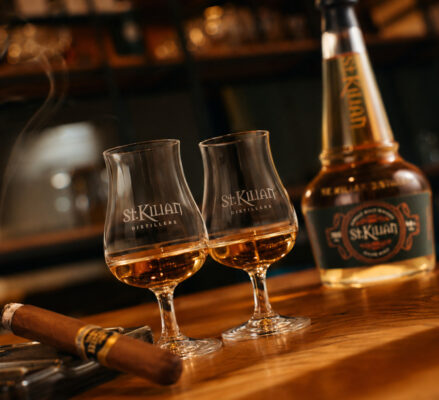 St. Kilian Cigar Malt zwei Gläser, links davon eine Zigarre, rechts im Hintergrund eine Flasche Whisky