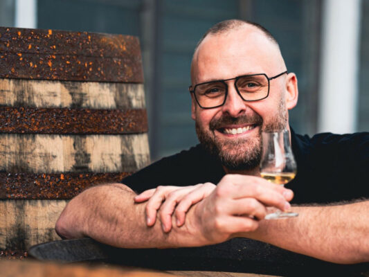 Mario Rudolf das Foto zeigt Mario Rudolf mit einem Glas Whisky, lehnend über ein Holzfass