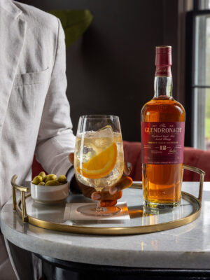 Spanish Highball der Drink "Spanish Highball" nebst einer Flasche The Glendronach 12 Jahre und einer Schale Oliven auf einem Tablett. Ein Mann im Sakko greift zum Drink