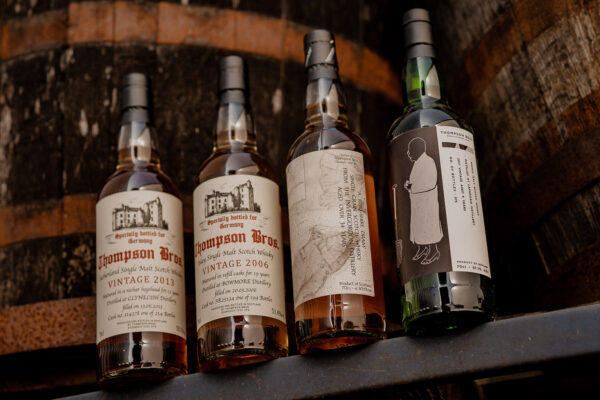 Thompson Bros. Neuheiten das Foto zeigt vier Flaschen Whisky von Thompson Bros., aufgereiht auf einer Stange. Dahinter sieht man zwei Holzfässer