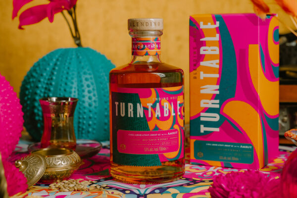 eine Flasche Turntable x Amrut Collaboration Drop 03 nebst Geschenkverpackung, umgeben von farbenfrohen Objekten mit indischem Touch