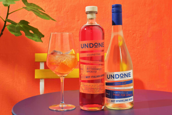 Undone das Foto zeigt zwei Flaschen und ein Glas auf einem Gartentisch. Im Hintergrund eine orangene Wand