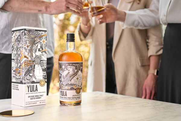 Yula Speyside Single Malt eine Flasche Yula Speyside Single Malt nebst Geschenkverpackung steht auf einem Tisch im Freien. Im Hintergrund mehrere Menschen, die anstoßen