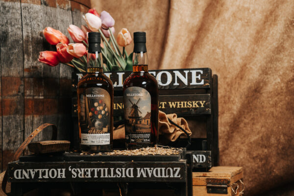 Zuidam Millstone 2019/2026 Banyuls Hogsheads und Zuidam Millstone 2020/2026 Heavily Peated Sauternes Hogsheads zwei Flaschen Whiskys auf einer Holzkiste, drum herum weitere Holzkisten und ein Holzfass